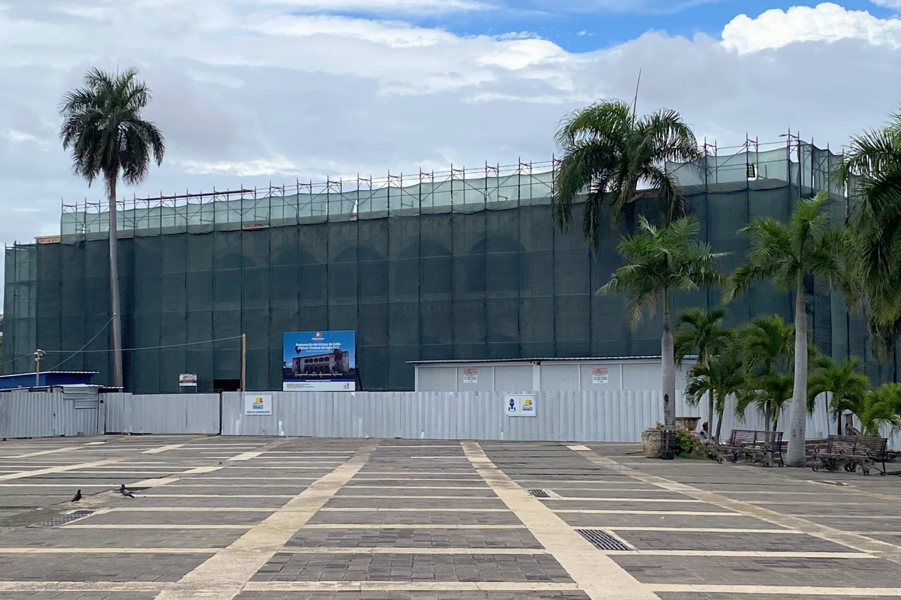 Edificio colonial cubierto con andamios y malla verde de obra, detrás de una verja, frente a una plaza con palmas